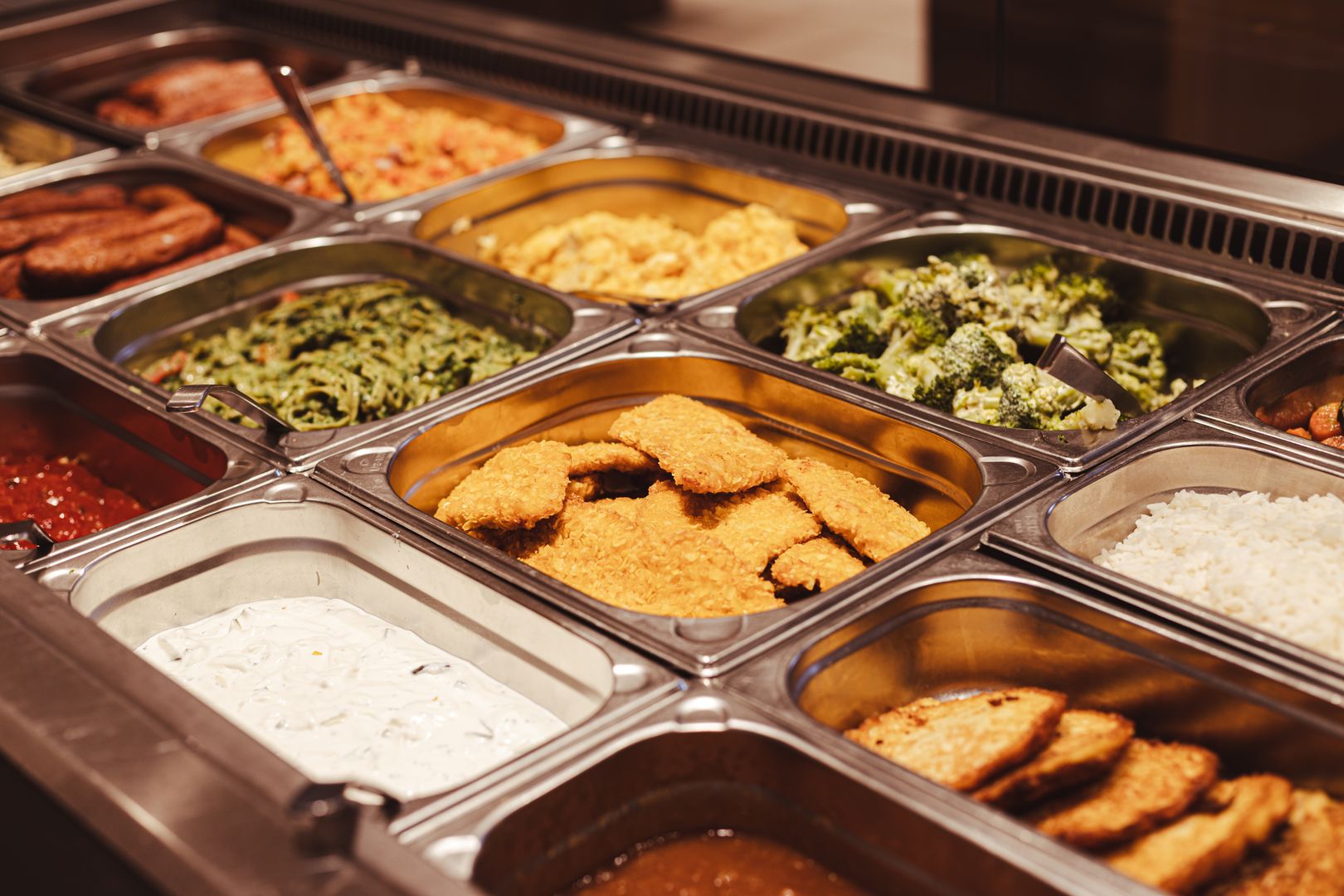 Buffet mit verschiedenen warmen und kalten Speisen in Edelstahlbehältern, darunter paniertes Schnitzel, Gemüse, Soßen und Salate